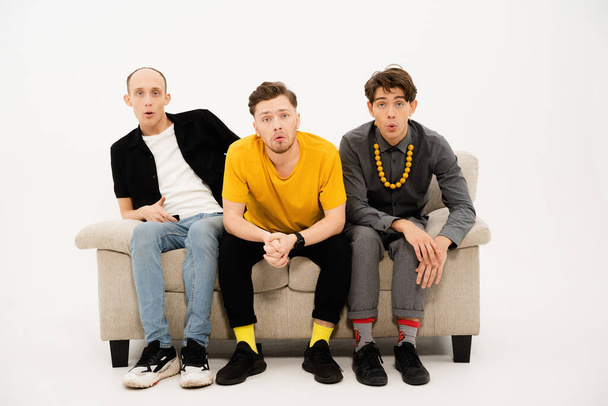 Portrait of three young friends spend time together sitting on the small sofa looking at the camera isolated on white background. Group of guys sitting on couch and communicates on white - Foto, Imagen