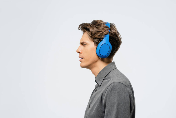Portrait of a young guy in grey outfit standing sideways listening music wearing blue wireless headphones. Funny young guy listen to his favourite track or song - Foto, imagen