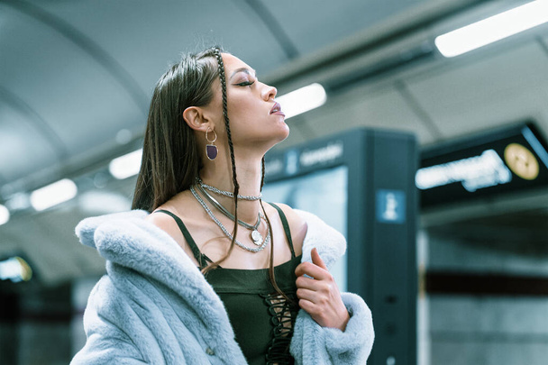 Fashion model young woman in purple synthetic fur coat bared her shoulders walking in metro or subway platform. Female with luxury makeup, long hair in stylish outfit - Photo, Image