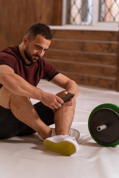 Handsome man sitting on a floor with smartphone in his hands and black and green tone fitness barbell, equipment for weight training concept. Sports equipment for training. Healthy lifestyle concept - Фото, изображение