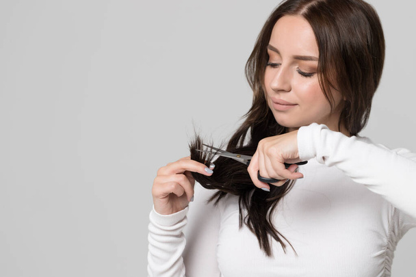 Trim hair, separated dry ends, hair care problems. Closeup portrait of brunette woman looking and cutting her split ends, holding scissors, isolated - Photo, Image