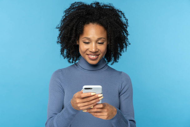 Felice donna africana millenaria con capelli ricci che chatta sui social media, utilizzando il telefono cellulare isolato - Foto, immagini