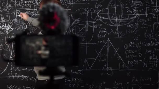 Um jovem professor dá uma lição de matemática on-line na frente de uma câmera de smartphone em pé no quadro - Filmagem, Vídeo