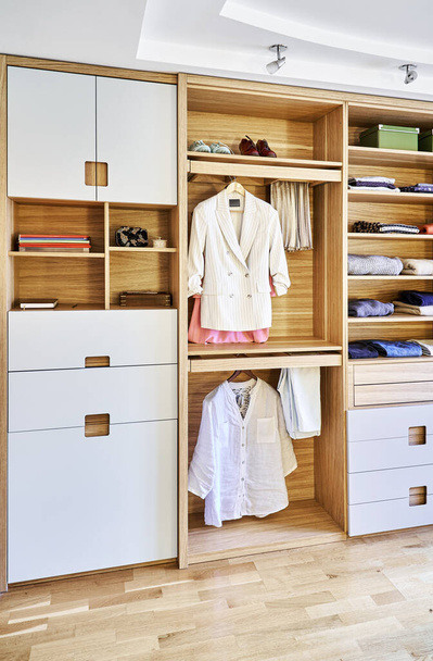 Internal details of the wooden wardrobe with slide out rack for coathangers. Modern wardrobe with clothes hanging on slide out racks and folded on the shelves. Modern furniture - 写真・画像