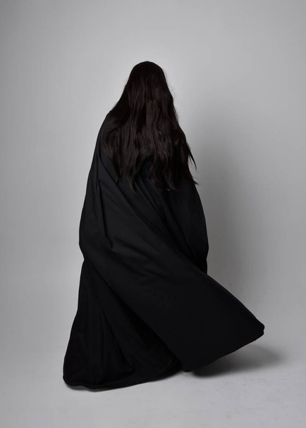 Full length portrait of pretty black haired woman wearing long dark gown nada cloak.  Standing pose facing away from the camera, against a  studio background. - Fotó, kép
