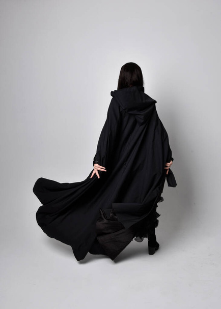 Full length portrait of pretty black haired woman wearing long dark gown nada cloak.  Standing pose facing away from the camera, against a  studio background. - Foto, Bild