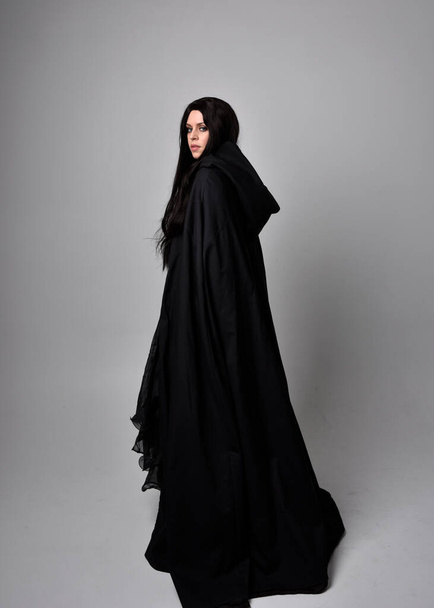Retrato de cuerpo entero de una bonita mujer de pelo negro con un largo vestido oscuro nada capa. Posar de pie frente a la cámara, sobre un fondo de estudio. - Foto, Imagen