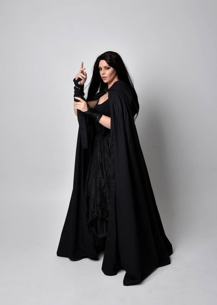 Full length portrait of pretty black haired woman wearing long dark gown nada cloak.  Standing pose facing away from the camera, against a  studio background. - Fotó, kép