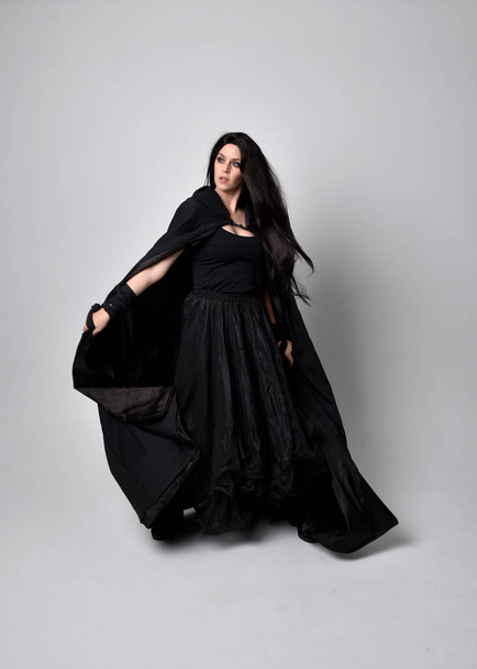 Full length portret van mooie zwart harige vrouw met lange donkere jurk nada mantel. Staande houding tegenover de camera, tegen een studio achtergrond. - Foto, afbeelding