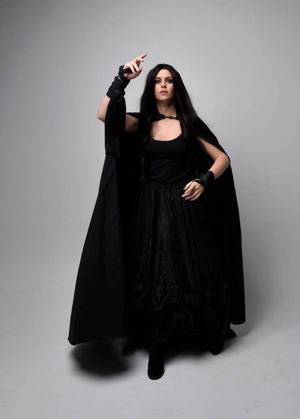 Full length portrait of pretty black haired woman wearing long dark gown nada cloak.  Standing pose facing away from the camera, against a  studio background. - Zdjęcie, obraz