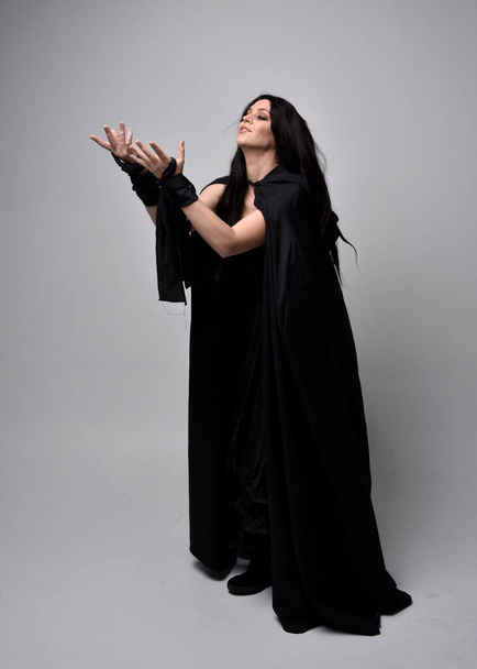 Full length portrait of pretty black haired woman wearing long dark gown nada cloak.  Standing pose facing away from the camera, against a  studio background. - Photo, Image