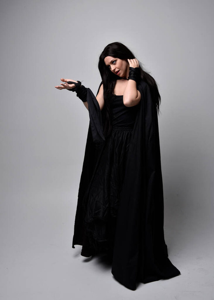 Full length portrait of pretty black haired woman wearing long dark gown nada cloak.  Standing pose facing away from the camera, against a  studio background. - Foto, Imagem