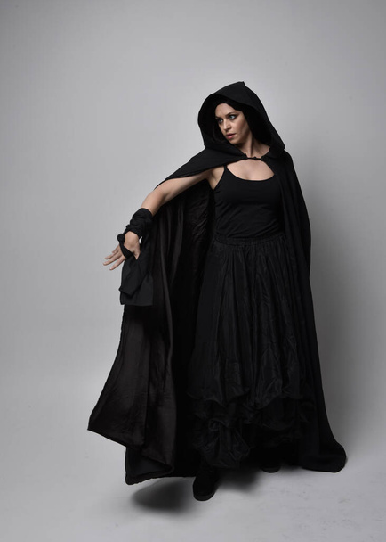 Retrato de comprimento total de mulher de cabelos pretos bonita vestindo manto longo vestido escuro nada. Posição em pé de frente para longe da câmera, contra um fundo de estúdio. - Foto, Imagem
