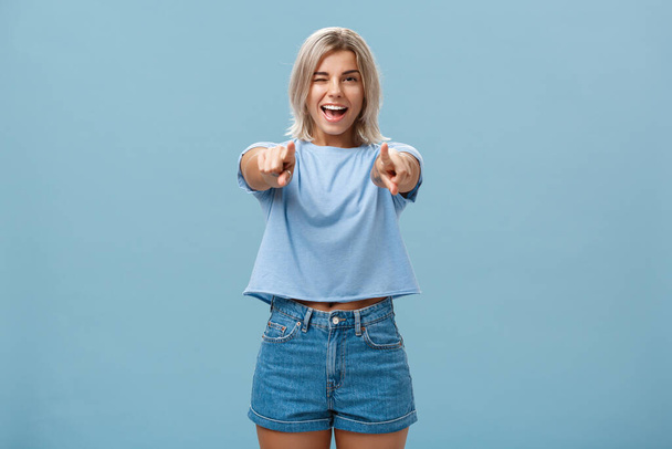 Glücklich enthusiastische, stylische blonde Frau mit Tätowierungen, die in die Kamera zeigen, als ob sie freudig lächelnd und zwinkernd vor Erstaunen und Glück vor blauem Hintergrund posiert - Foto, Bild