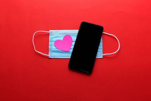 Ημέρα του Αγίου Βαλεντίνου σε πανδημία από covid-19, κινητό τηλέφωνο με μάσκα προστασίας κλινική χρήση και ροζ καρδιές - Φωτογραφία, εικόνα
