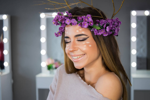 Jovem mulher com chifres de ramos de coroa de flores violeta e pontos de veado no rosto de pé olhando para a câmera sorrindo no fundo espelhos - Foto, Imagem
