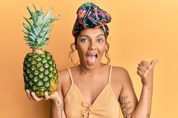 Young hispanic girl wearing hair turban holding pineapple pointing thumb up to the side smiling happy with open mouth  - Foto, immagini