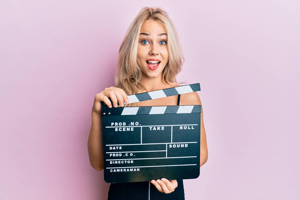 Beautiful caucasian blonde girl holding video film clapboard celebrating crazy and amazed for success with open eyes screaming excited.  - Foto, afbeelding