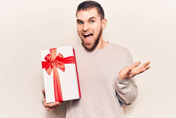 Young handsome man holding gift celebrating achievement with happy smile and winner expression with raised hand  - Photo, Image