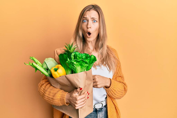Beautiful caucasian woman holding paper bag with bread and groceries scared and amazed with open mouth for surprise, disbelief face  - Photo, Image