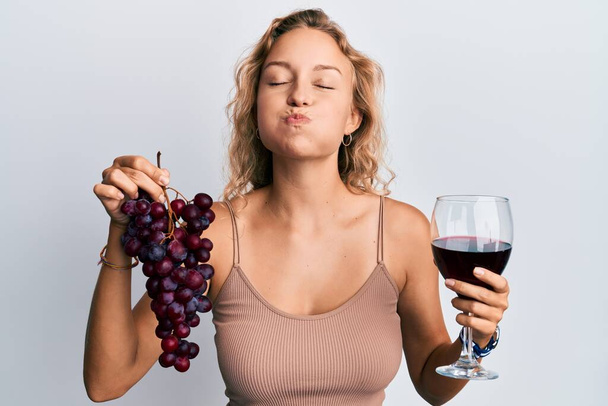 Beautiful caucasian woman holding branch of fresh grapes and red wine puffing cheeks with funny face. mouth inflated with air, catching air.  - Zdjęcie, obraz