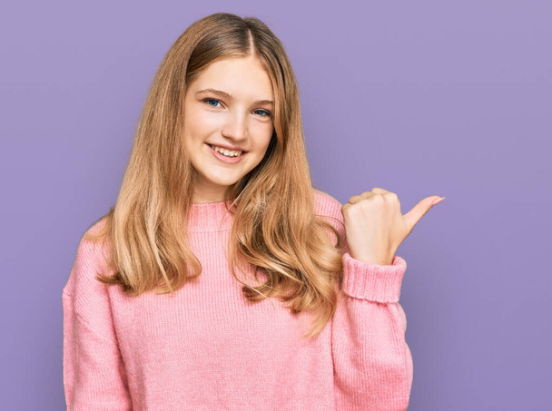 Beautiful young caucasian girl wearing casual winter sweater smiling with happy face looking and pointing to the side with thumb up.  - 写真・画像