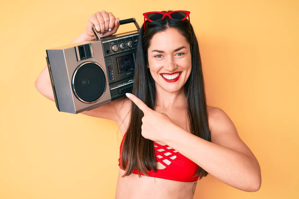 Jonge mooie blanke vrouw draagt bikini en houdt boombox glimlachend gelukkig wijzend met hand en vinger  - Foto, afbeelding
