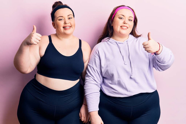 Young plus size twins wearing sportswear doing happy thumbs up gesture with hand. approving expression looking at the camera showing success.  - Fotografie, Obrázek