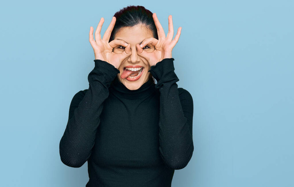Mujer hispana joven vestida con ropa casual haciendo un buen gesto como binoculares sacando la lengua, ojos mirando a través de los dedos. expresión loca.  - Foto, imagen