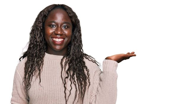 Mladá africká žena na sobě vlny zimní svetr s úsměvem veselý prezentaci a ukazuje s dlaní ruky při pohledu na fotoaparát.  - Fotografie, Obrázek