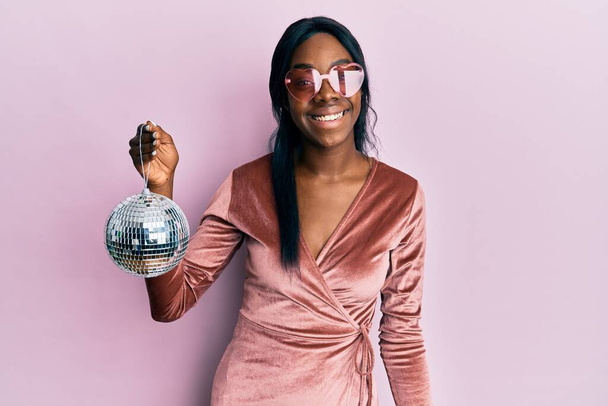 Joven mujer afroamericana vistiendo vestido de fiesta sexy sosteniendo la bola disco mirando positiva y feliz de pie y sonriendo con una sonrisa confiada mostrando los dientes  - Foto, imagen