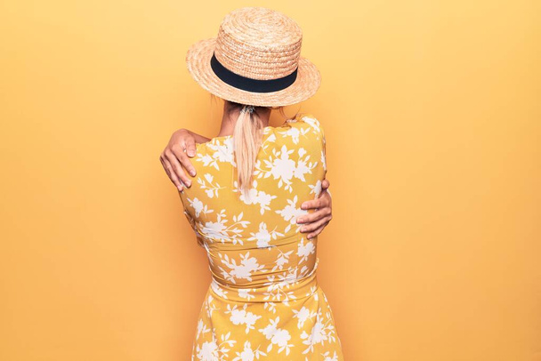 Hermosa mujer rubia de vacaciones con sombrero de verano y vestido sobre fondo amarillo Abrazarse feliz y positivo desde atrás. Amor propio y cuidado personal - Foto, Imagen