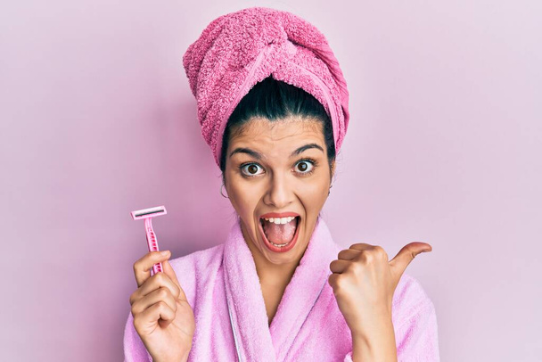 Nuori latino nainen yllään suihku kylpytakki tilalla partaveitsi osoittaa peukalo ylös puolelle hymyillen onnellinen suu auki  - Valokuva, kuva