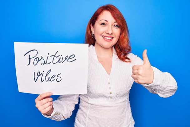 Young redhead woman asking for optimist attitude holding paper with positive vibes message smiling happy and positive, thumb up doing excellent and approval sign - Photo, Image