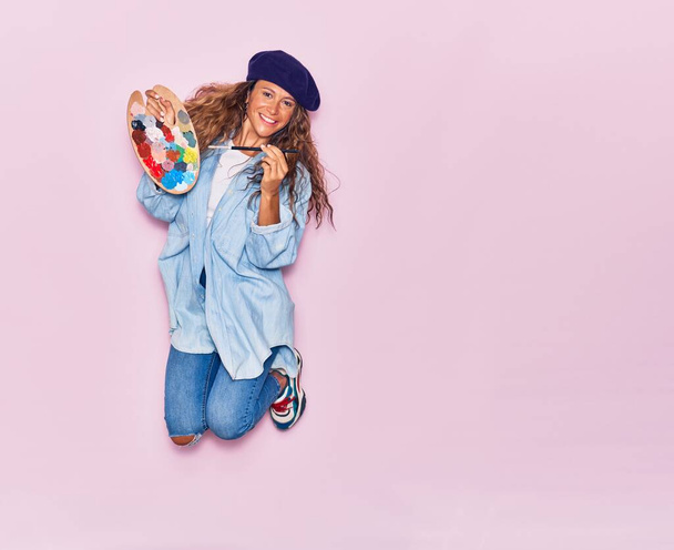 Young beautiful curly artist woman with tattoo wearing french beret smiling happy. Jumping with smile on face painting using palette and paintbrush over isolated pink background - Photo, image