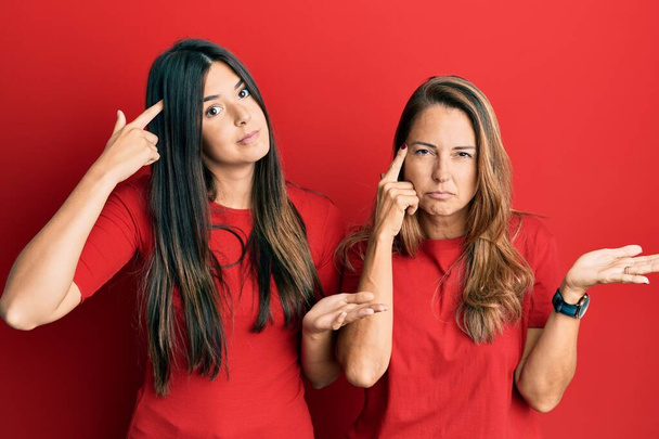 赤い背景の上にカジュアルな服を着ている母親と娘のヒスパニック系の家族は混乱し、コピースペースを示し、額に指を指すオープンヤシに悩まされています。考えてみてください.  - 写真・画像