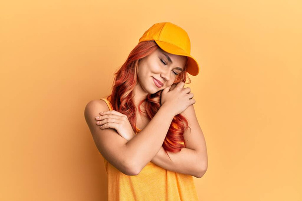 Jonge mooie roodharige vrouw met casual kleding en gele pet knuffelend zichzelf gelukkig en positief, glimlachend vol vertrouwen. zelfliefde en zelfzorg  - Foto, afbeelding