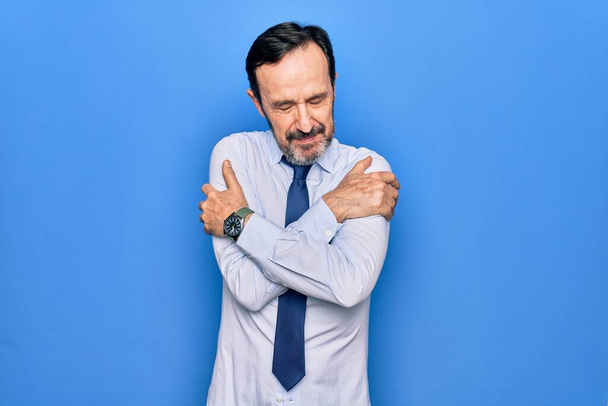 Keski-ikä komea liikemies yllään tyylikäs solmio seisoo eristetty sininen tausta halaamalla itseään onnellinen ja positiivinen, hymyillen luottavainen. Itserakkaus ja itsehoito - Valokuva, kuva