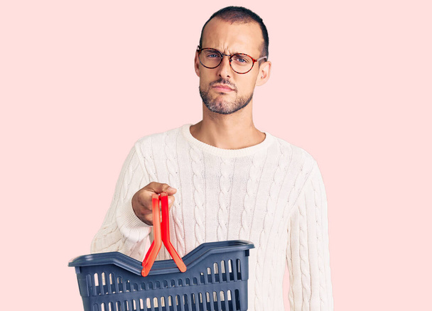 Young handsome man holding supermarket shopping basket thinking attitude and sober expression looking self confident  - Photo, Image