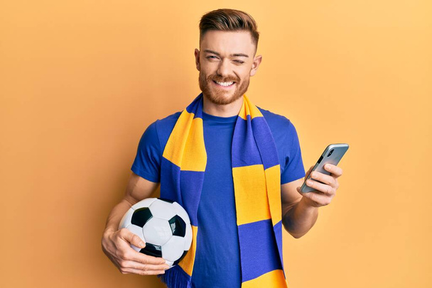 Young redhead man wearing hooligan scarf and using smartphone winking looking at the camera with sexy expression, cheerful and happy face.  - 写真・画像