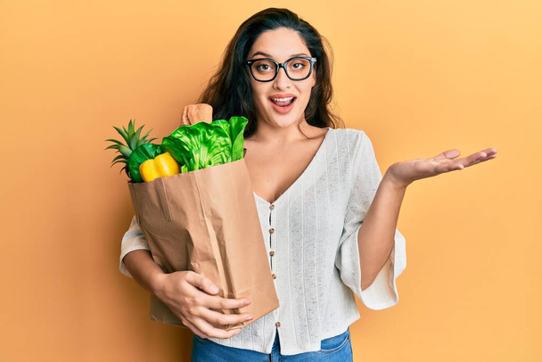Beautiful middle eastern woman holding paper bag with groceries celebrating achievement with happy smile and winner expression with raised hand  - Photo, Image
