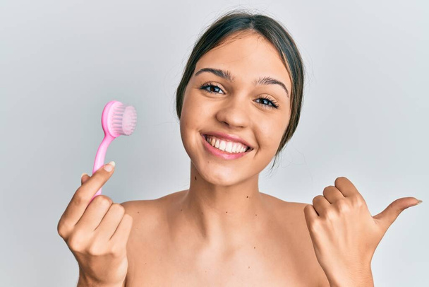 Young brunette woman using facial exfoliating brush pointing thumb up to the side smiling happy with open mouth  - Foto, imagen