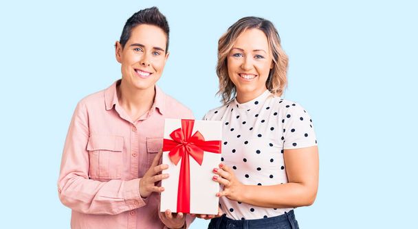 Пара женщин, держащих в руках подарок выглядит позитивно и счастливо стоя и улыбаясь с уверенной улыбкой, показывая зубы  - Фото, изображение