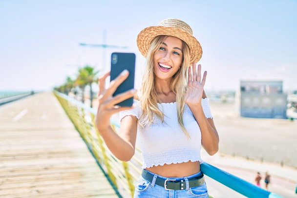 Giovane bionda turista sorridente felice facendo videochiamata utilizzando smartphone sul lungomare. - Foto, immagini