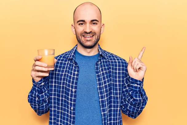Fiatal, jóképű, kopasz férfi, egy pohár egészséges narancslevet iszik, mosolyogva, boldogan mutatva kezével és ujjával oldalra.  - Fotó, kép