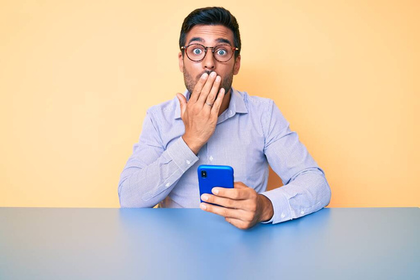 Jonge Spaanse man met behulp van smartphone zitten op de tafel bedekken mond met de hand, geschokt en bang voor fouten. verraste uitdrukking  - Foto, afbeelding