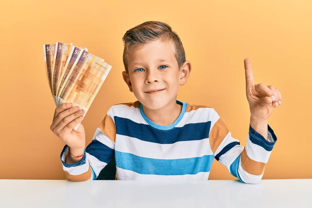 Urocze dziecko rasy kaukaskiej trzymające 500 norweskich banknotów koronowych siedzących na stole i uśmiechających się z pomysłem lub pytaniem wskazującym palcem szczęśliwą twarz, numer jeden  - Zdjęcie, obraz