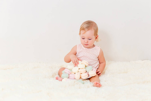 Cute European baby girl one year old sits on a white fluffy plaid and plays and marshmallows on white background - Foto, Imagem