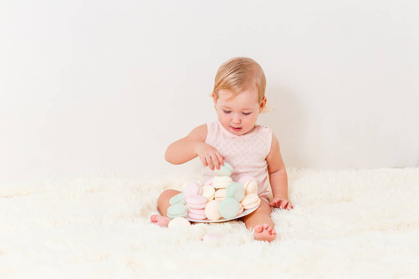Cute European baby girl one year old sits on a white fluffy plaid and plays and marshmallows on white background - Foto, Imagem