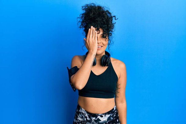 Young hispanic woman wearing gym clothes and using headphones covering one eye with hand, confident smile on face and surprise emotion.  - Foto, afbeelding